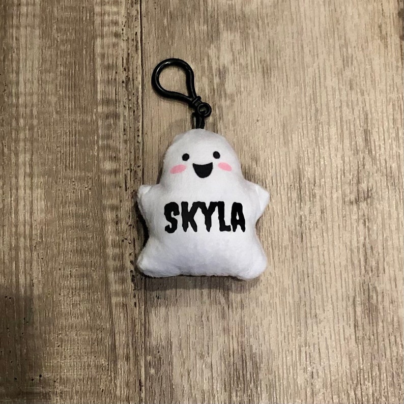 May include: White plush ghost keychain with a black clip. The ghost has a smiling face with pink cheeks and the name "Skyla" written in black.