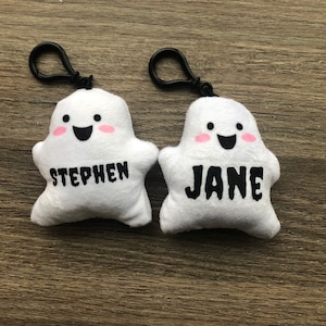 May include: Two white plush ghost keychains with black eyes and mouths. The ghost on the left has the name "STEPHEN" embroidered on it, and the ghost on the right has the name "JANE" embroidered on it.