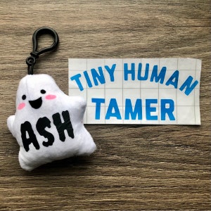 May include: White plush ghost keychain with a smiling face and the name "Ash" written in black. The keychain is attached to a white sticker with the text "Tiny Human Tamer" in blue.