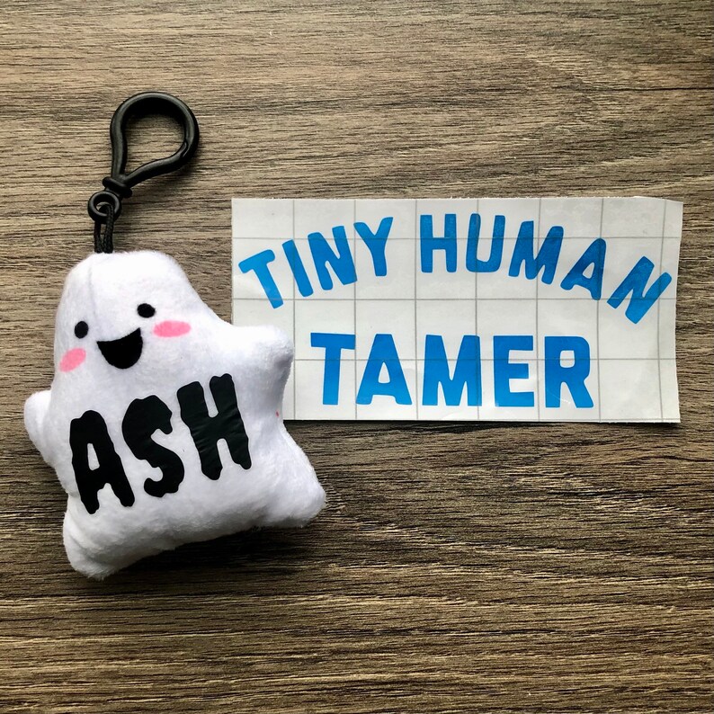 May include: White plush ghost keychain with a smiling face and the name "Ash" written in black. The keychain is attached to a white sticker with the text "Tiny Human Tamer" in blue.