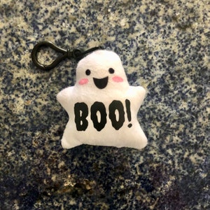 May include: White plush ghost keychain with a black "BOO!" on the front and a black clip. The ghost has a smiling face with pink cheeks.