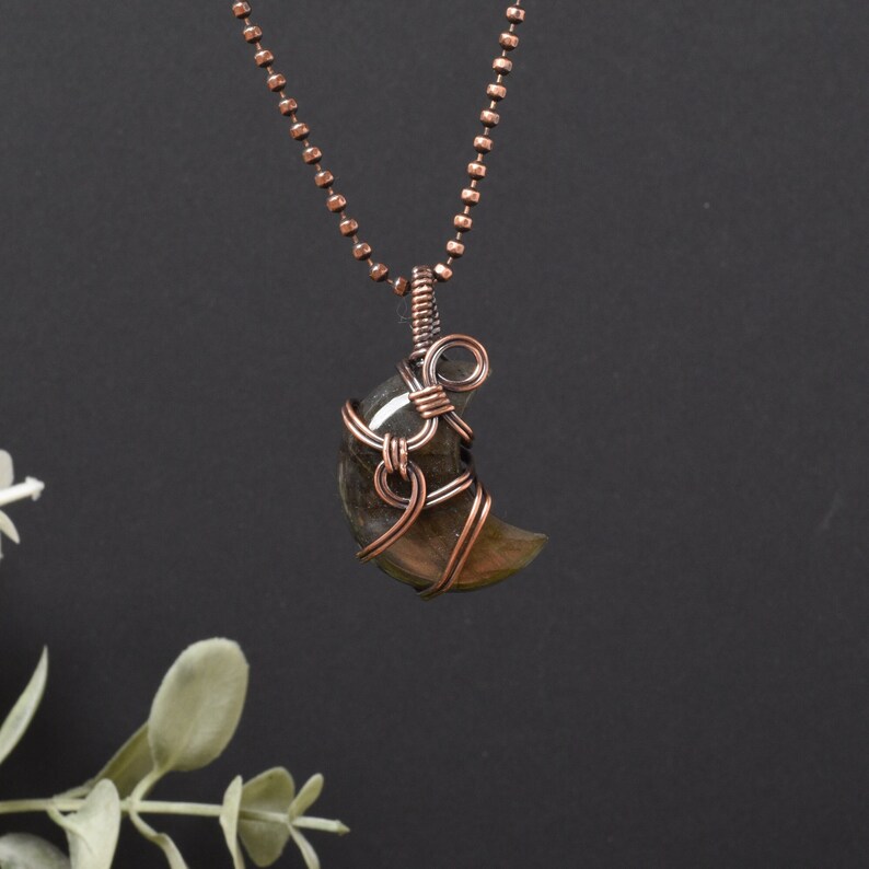 A beautiful golden crescent moon labradorite delicately wrapped in antiqued copper wire suspended from a copper ball chain against a black background by Lyn Haslegrave Jewellery