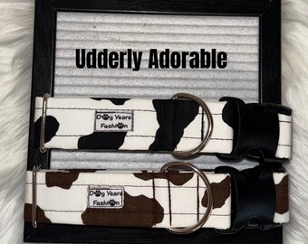 May include: Two dog collars with a cow print design, one in black and white, the other in brown and white. Each collar has a black plastic buckle and a silver metal D-ring. The collars are displayed on a black framed sign that reads "Udderly Adorable."