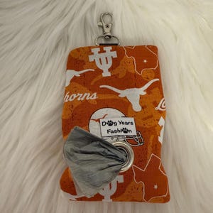 May include: Orange and white patterned dog waste bag dispenser with a silver carabiner clip. The fabric features the word "horns", a longhorn logo, and a map of Texas. The dispenser has a metal grommet and a roll of gray waste bags. The text "Dog Years Fashion" is visible.