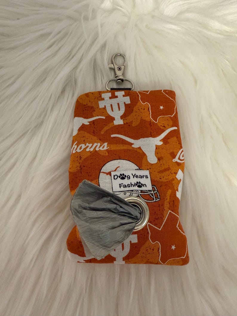 May include: Orange and white patterned dog waste bag dispenser with a silver carabiner clip. The fabric features the word "horns", a longhorn logo, and a map of Texas. The dispenser has a metal grommet and a roll of gray waste bags. The text "Dog Years Fashion" is visible.