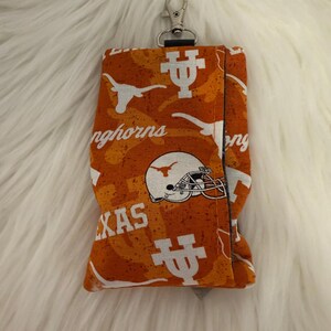 May include: Orange and white fabric key fob holder with a silver clasp. The fabric features a repeating pattern of the Texas Longhorns logo, including the Longhorn silhouette, a football helmet, and the word "Texas".