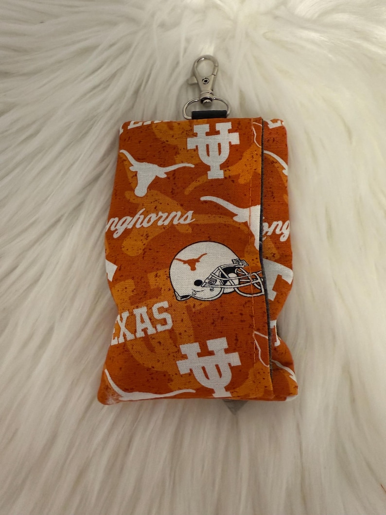 May include: Orange and white fabric key fob holder with a silver clasp. The fabric features a repeating pattern of the Texas Longhorns logo, including the Longhorn silhouette, a football helmet, and the word "Texas".