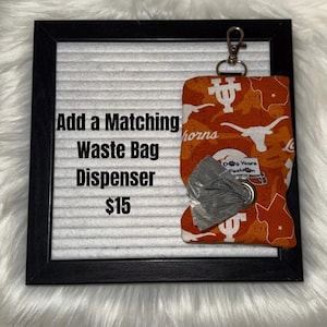May include: Orange and white patterned waste bag dispenser with a silver clip. The dispenser is attached to a black framed board with the text "Add a Matching Waste Bag Dispenser". The dispenser has a roll of gray waste bags.