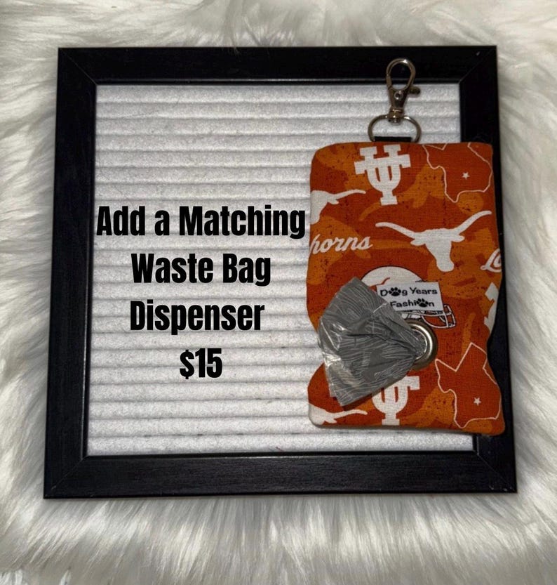 May include: Orange and white patterned waste bag dispenser with a silver clip. The dispenser is attached to a black framed board with the text "Add a Matching Waste Bag Dispenser". The dispenser has a roll of gray waste bags.