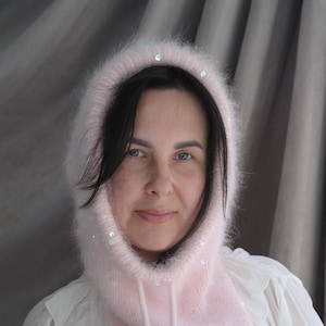 May include: A light pink knitted hood with a drawstring and embellished with sequins. The hood is made of a soft, fuzzy material and is designed to be worn around the head and neck. The hood is a stylish accessory.