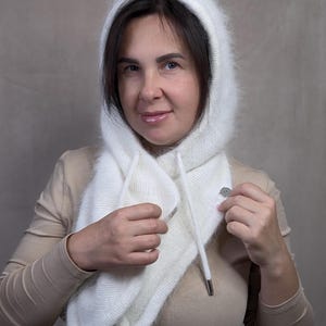 May include: A white fuzzy hooded scarf featuring a soft, brushed texture. The hood is integrated into a scarf design that wraps around the neck, secured with a silver-toned metal snap button. Two long white drawstrings with silver metal tips hang down.