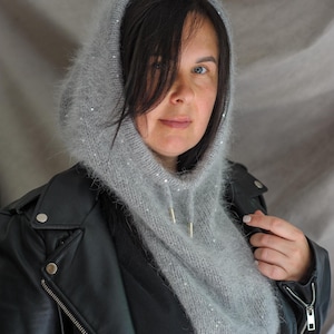 Peut inclure: Une cagoule grise tricotée avec un cordon de serrage et des paillettes scintillantes. La cagoule est faite d'une matière douce et duveteuse et se porte sur la tête et autour du cou. La personne sur la photo porte une veste en cuir noire.