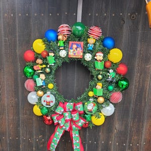May include: A festive Christmas wreath with a green base, adorned with colorful ornaments, Snoopy-themed figurines, and a red and green bow. The wreath features a variety of ball ornaments in red, yellow, green, and blue, creating a cheerful holiday display.