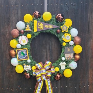 May include: A festive wreath featuring a baseball theme. It includes a variety of decorative elements such as yellow and brown ornaments, a Padres pennant, and a large bow. The wreath is adorned with baseball-related accents.