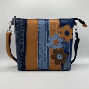 May include: A denim crossbody bag with a patchwork design. The bag features vertical stripes of blue denim and tan fabric. Three appliqued flower shapes in blue and brown adorn the front. The bag has a brown strap and silver hardware.
