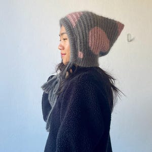 Può includere: Un cappuccio lavorato a maglia grigio e rosa con paraorecchie e una sciarpa abbinata. Il cappuccio presenta un design a coste con accenti rosa. La sciarpa è in maglia grigia simile. Abbigliamento fatto a mano, adatto al freddo.