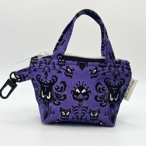 May include: A small purple fabric pouch with two handles and a white zipper closure. The fabric features a repeating black pattern of stylized faces with white eyes. A black plastic clip is attached to the side, and a small white tag reads 4th and Leo.