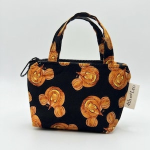May include: A small black fabric pouch with two handles and a zipper closure, featuring an all-over print of orange pumpkin shapes resembling mouse ears. A white fabric tag on the side displays the text 4th and Leo in black lettering.
