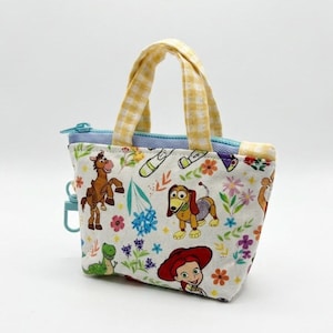 May include: A small fabric pouch with two yellow and white gingham handles. The white fabric features a colorful print of cartoon characters including a brown horse, a dachshund, and a red-haired figure, alongside green and blue floral accents.