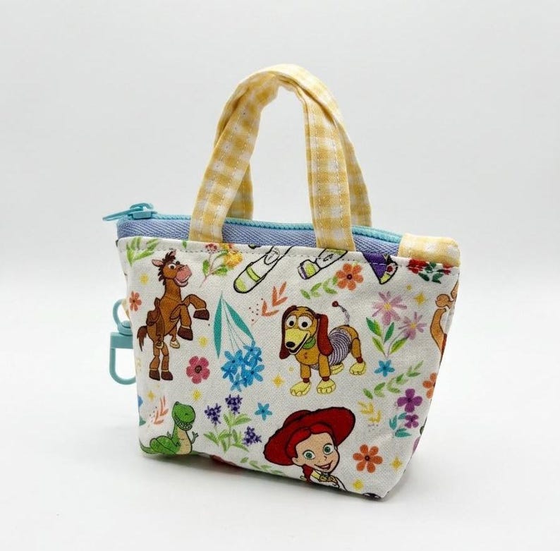 May include: A small fabric pouch with two yellow and white gingham handles. The white fabric features a colorful print of cartoon characters including a brown horse, a dachshund, and a red-haired figure, alongside green and blue floral accents.