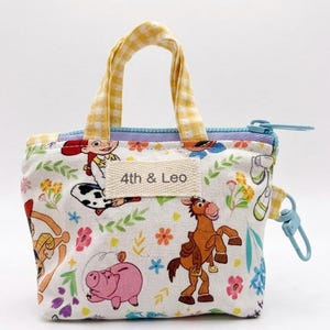 May include: A small fabric pouch with yellow gingham handles and a light blue zipper. The white exterior features a colorful print of cartoon characters, flowers, and greenery. A rectangular fabric label on the front displays the text 4th & Leo.