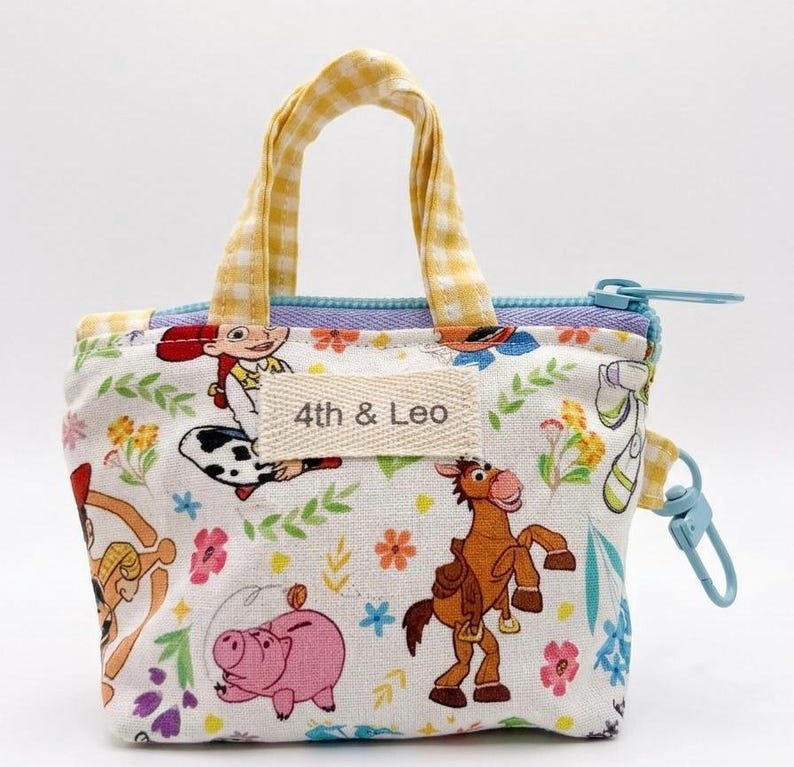 May include: A small fabric pouch with yellow gingham handles and a light blue zipper. The white exterior features a colorful print of cartoon characters, flowers, and greenery. A rectangular fabric label on the front displays the text 4th & Leo.
