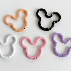 May include: Five circular spring-loaded carabiner clips shaped like mouse heads. The set includes one peach, one black, one white, one light pink, and one lavender clip. Each metal ring features a hinged opening mechanism for attaching items.