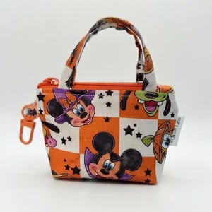 Könnte beinhalten: Ein kleines Stofftäschchen mit orange-weißem Karomuster und Cartoon-Figuren in Halloween-Kostümen, darunter eine Vampir-Maus und eine Hexen-Maus. Das Täschchen verfügt über einen orangefarbenen Reißverschluss, zwei Tragegriffe und einen Clip.