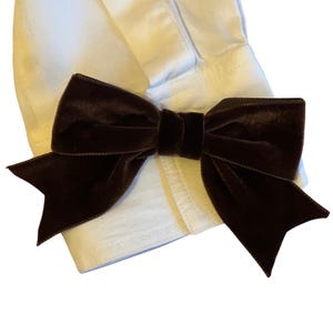 May include: A dark brown velvet bow tie is attached to a white shirt cuff. The bow tie is centered and has a classic bow shape. The shirt cuff is white and has a buttoned closure.