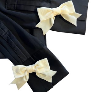 May include: Black garment with cream-colored velvet bows. The bows are tied in the center and have a classic bow shape. The garment appears to be a jacket or top, with details like pleats and cuffs visible.