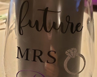 May include: Clear glass wine glass with the text "future MRS Pina" in black and purple lettering. A silver glitter ring with a diamond is next to the text.