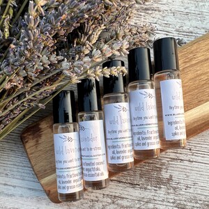 May include: Five glass roll-on bottles with black caps, labeled "wild lavender" and "lavender", are arranged on a wooden surface with dried lavender sprigs. The bottles have white labels with text and a lavender sprig illustration.