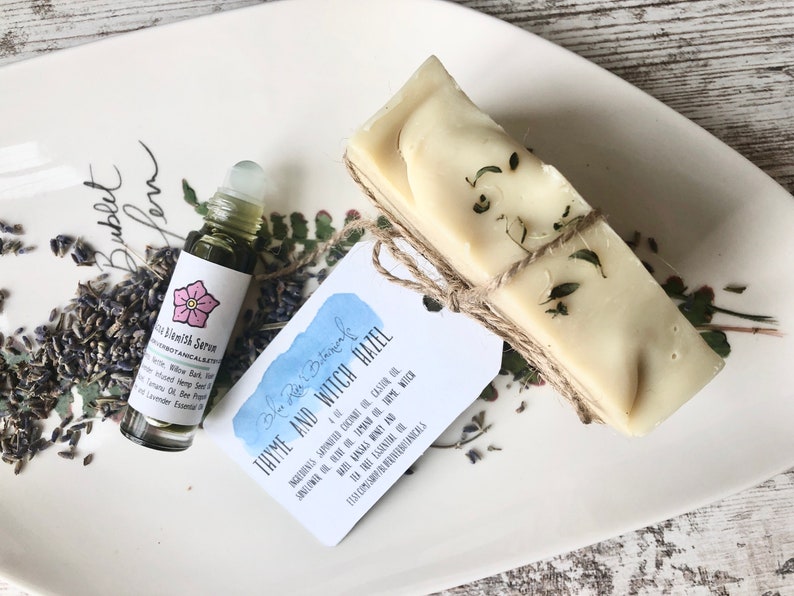 May include: A small bottle of serum with a pink flower graphic and the text "Blemish Serum" and a bar of soap wrapped in twine with a tag that reads "Blue Moon Botanicals Thyme and Witch Hazel" with a list of ingredients.