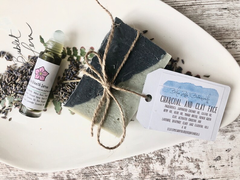 May include: A white ceramic plate with a bar of charcoal and clay face soap tied with twine, a small glass bottle of blemish serum, and dried lavender flowers. The soap label reads "Blue River Botanicals Charcoal and Clay Face".