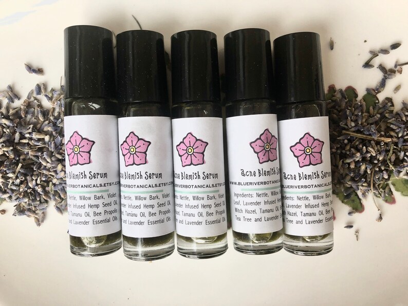 May include: Five small glass bottles with black roll-on tops and white labels. The labels have a pink flower graphic and the text "Acne Blemish Serum" with the website address "www.blueriverbotanicals.etsy.com". The bottles are on a white surface with dried lavender flowers scattered around them.