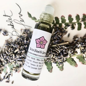 May include: A glass roller bottle with a green liquid and a white label that says "acne Blemish Serum" and "BLUERIVERBOTANICALSALES". The label also has a pink flower illustration. The bottle is surrounded by dried lavender buds.