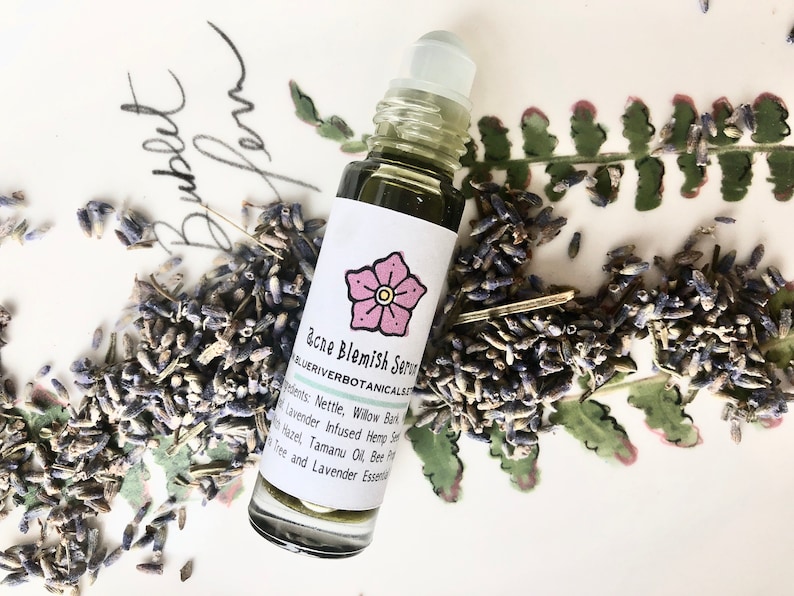 May include: A glass roller bottle with a green liquid and a white label that says "acne Blemish Serum" and "BLUERIVERBOTANICALSALES". The label also has a pink flower illustration. The bottle is surrounded by dried lavender buds.