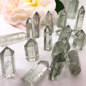 May include: A collection of clear green crystal points, also known as green quartz points, arranged on a white surface. The crystals are various sizes and shapes, with some having a more pointed tip than others.