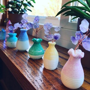 May include: A row of six small ceramic vases with a white base and a colored top. Each vase is topped with a small wire sculpture of amethyst crystals.