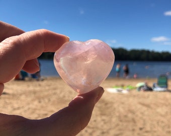 May include: A heart-shaped pink crystal, also known as rose quartz, is held in a hand against a backdrop of a sandy beach and blue sky.