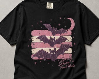 May include: A black short-sleeve t-shirt featuring a graphic of three purple bats arranged vertically over horizontal pink distressed stripes. A crescent moon and small stars surround the bats. The text "Spooky Season Feral Mode" is printed in pink.