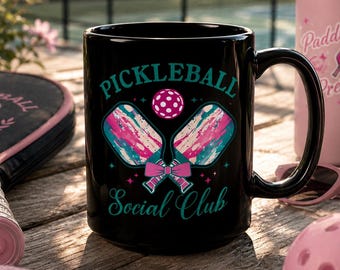 May include: A black ceramic mug featuring a graphic design with two crossed pickleball paddles in pink and teal, a pink pickleball, and a pink bow. The text reads PICKLEBALL SOCIAL CLUB in teal lettering, accented by small pink stars.