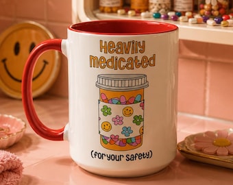 May include: A white ceramic mug with a red interior and handle, featuring a graphic of a pill bottle with a floral and smiley face pattern. The text reads "Heavily medicated" above the bottle and "(for your safety)" below the illustration.