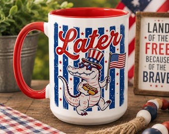May include: A white ceramic mug with a red interior and handle. The front features a graphic of an alligator wearing a patriotic top hat and star-shaped sunglasses, holding a hot dog and a small American flag, with the word Later printed above.