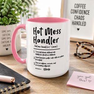 May include: A white ceramic mug with a pink interior and handle, featuring black text that reads Hot Mess Handler, a phonetic pronunciation, three numbered definitions, and the name Nicole printed at the bottom of the mug surface.