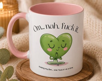 May include: A white ceramic mug with a soft pink interior and handle. The front features a green heart-shaped character in a meditation pose with the text "Om... nah, fuck it." and "Finding my Zen... one 'fuck it' at a time" printed in black.