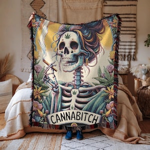 May include: A woven tapestry blanket featuring a skeleton with a cannabis leaf on its head, smoking a cigarette. The skeleton is surrounded by cannabis leaves and flowers, with the word "CANNABITCH" at the bottom. The blanket has a fringed edge.