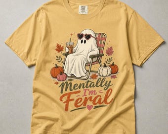 May include: A mustard yellow t-shirt featuring a graphic of a white ghost wearing heart-shaped sunglasses, sitting in a folding chair while holding a drink. The design includes pumpkins, autumn leaves, and the text Mentally I'm Feral.