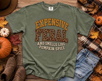 May include: An olive green short-sleeve t-shirt featuring the text EXPENSIVE in gold, FERAL in a brown leopard print pattern, and AND SMELLS LIKE PUMPKIN SPICE in white. The shirt has a crew neckline and a standard fit, displaying a casual design.