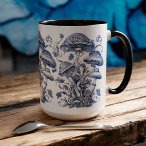 May include: A white ceramic mug with a black rim and handle features a blue and white illustration of mushrooms and flowers. The mug is sitting on a wooden surface.
